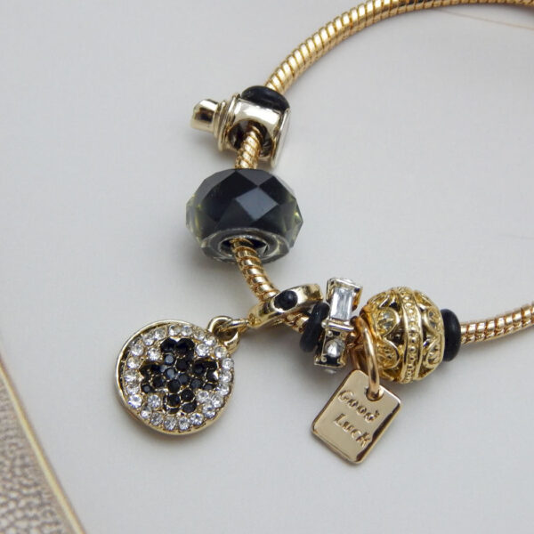 Gold-tone snake chain charm bracelet with black beads, ‘Good Luck’ tag, crystal accent charms on light background