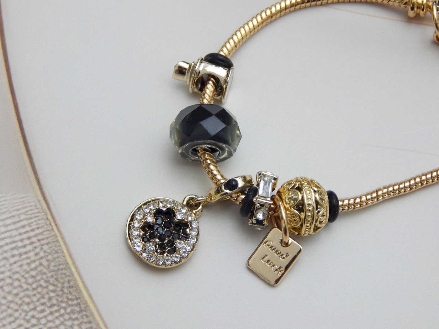Gold-tone snake chain charm bracelet with black beads, ‘Good Luck’ tag, crystal accent charms on light background