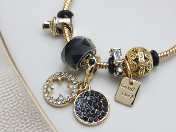 Gold-tone snake chain charm bracelet with black beads, ‘Good Luck’ tag, crystal accent charms on light background