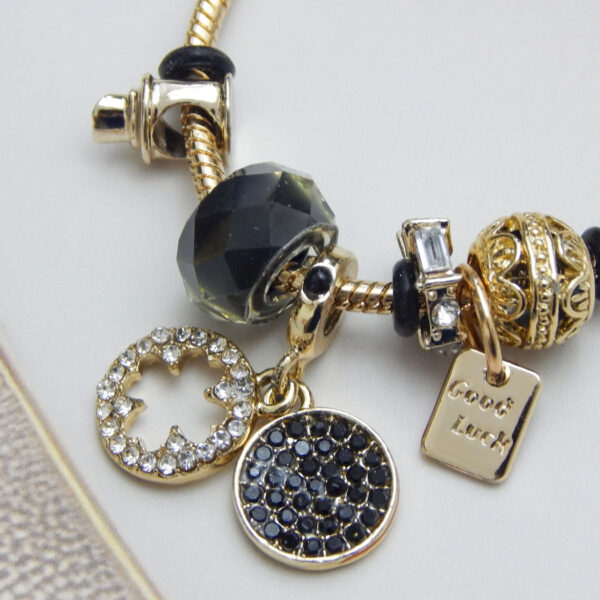 Gold-tone snake chain charm bracelet with black beads, ‘Good Luck’ tag, crystal accent charms on light background