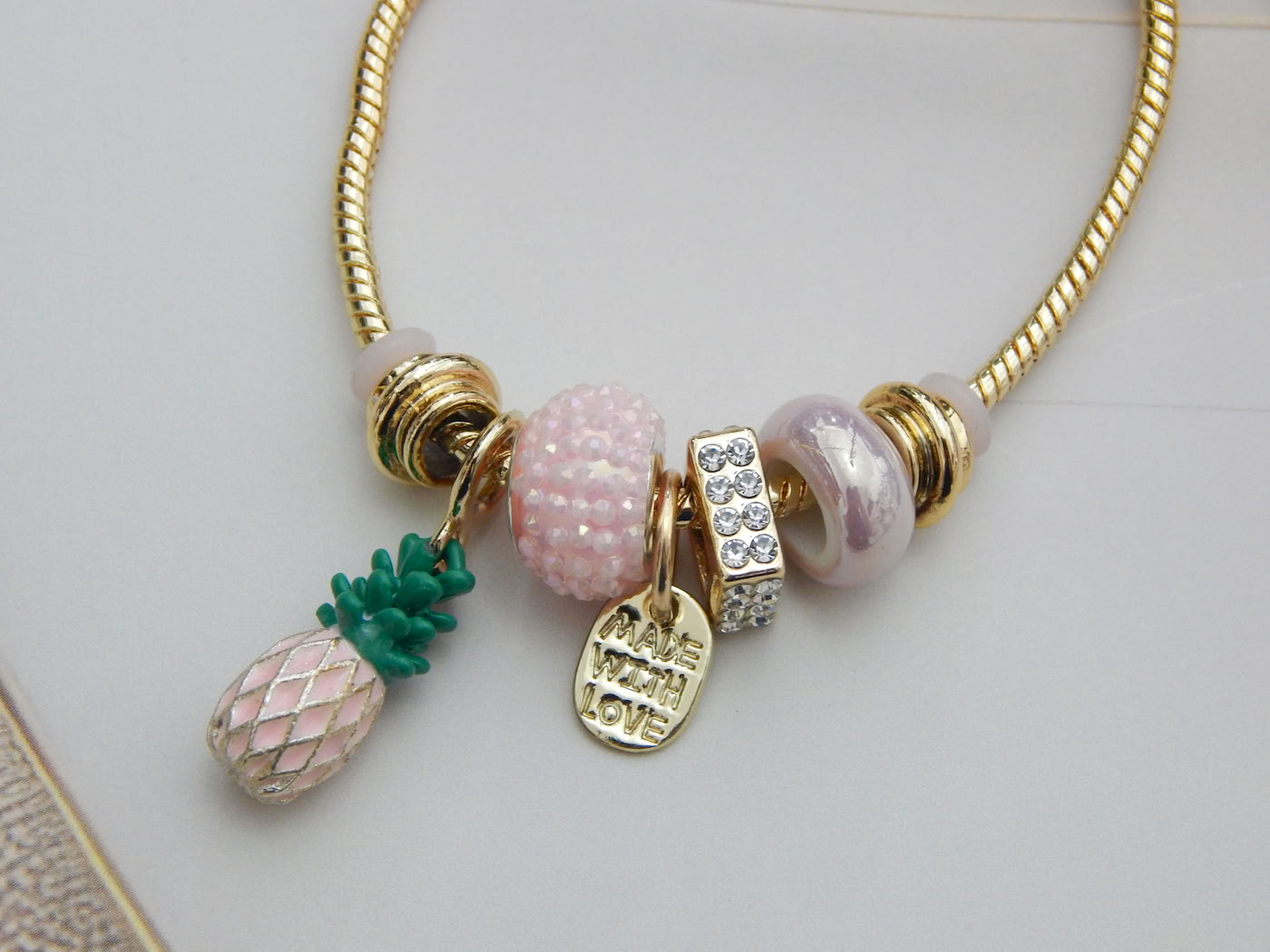 Bracelet with pink enamel pineapple charm, crystal spacer, Murano‑style beads, “Made With Love” pendant on gold snake chain
