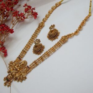 A traditional Indian temple jewelry necklace and earring set with an intricate Laxmi goddess pendant and floral designs, featuring ruby stones and cubic zirconia with a matte gold finish.