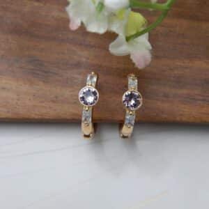 A pair of small, gold-toned huggie hoop earrings with a central round amethyst-colored stone and rectangular baguette-cut stones on the sides.