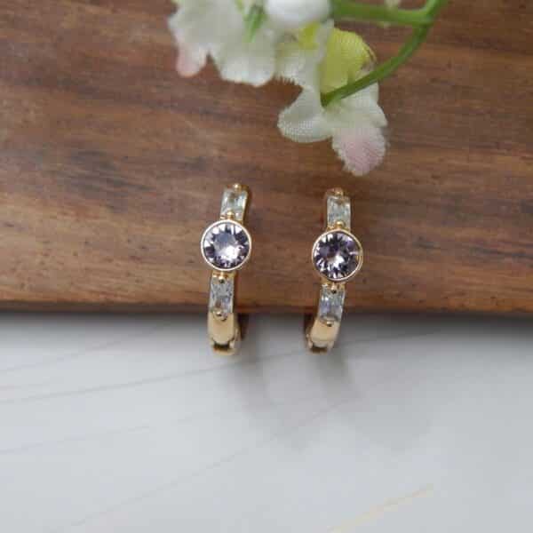 A pair of small, gold-toned huggie hoop earrings with a central round amethyst-colored stone and rectangular baguette-cut stones on the sides.