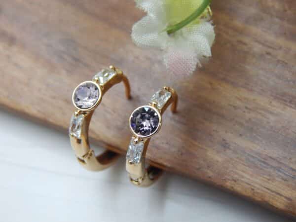 A pair of small, gold-toned huggie hoop earrings with a central round amethyst-colored stone and rectangular baguette-cut stones on the sides.