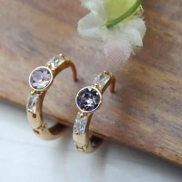 A pair of small, gold-toned huggie hoop earrings with a central round amethyst-colored stone and rectangular baguette-cut stones on the sides.