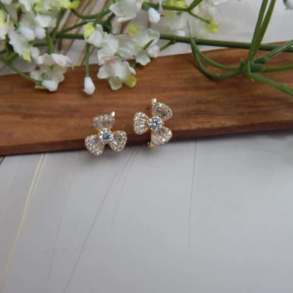 A pair of small, gold-toned huggie hoop earrings with a bow design, featuring a central round stone and baguette-cut stones.