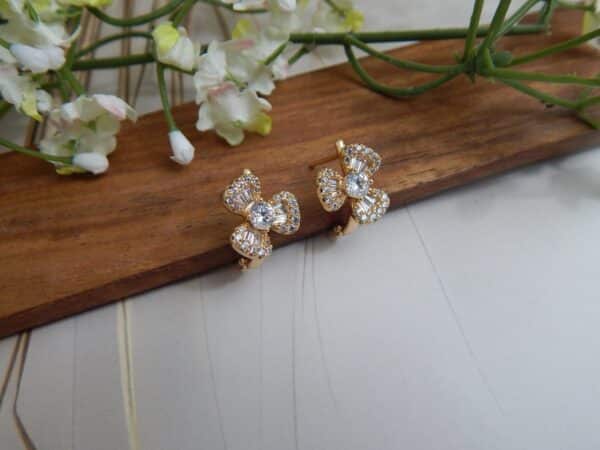 A pair of small, gold-toned huggie hoop earrings with a bow design, featuring a central round stone and baguette-cut stones.