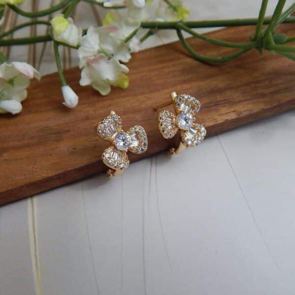 A pair of small, gold-toned huggie hoop earrings with a bow design, featuring a central round stone and baguette-cut stones.