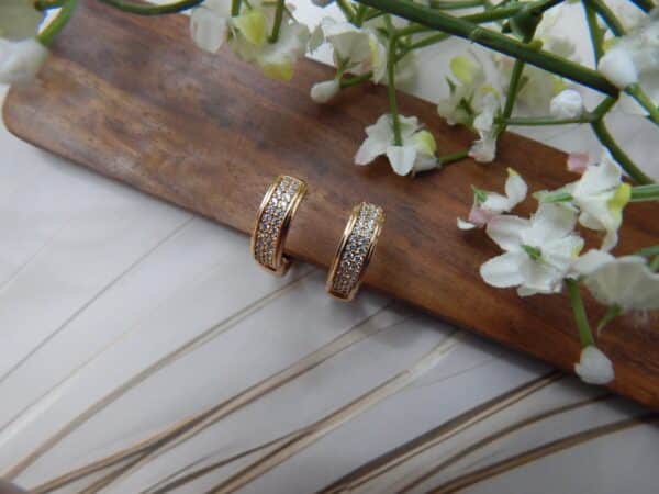 A pair of small, gold-toned huggie hoop earrings with a rectangular shape and rows of clear studs