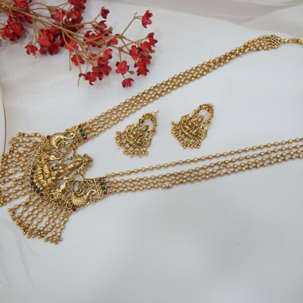 A traditional long Indian temple jewelry necklace featuring a Laxmi goddess pendant with peacock motifs, intricate beaded chains, and a matte gold finish.