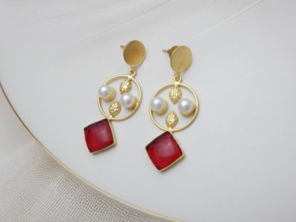 A pair of gold-tone geometric dangle earrings with a circle frame, white pearls, and a large square-shaped red stone.
