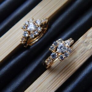 A pair of small, gold-toned huggie hoop earrings featuring a central heart-shaped stone surrounded by clear studs.