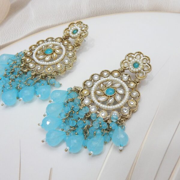A pair of large, traditional dangle earrings with a central oval stone, intricate work, a halo of small pearls, and a fan of dangling light blue colored beads.