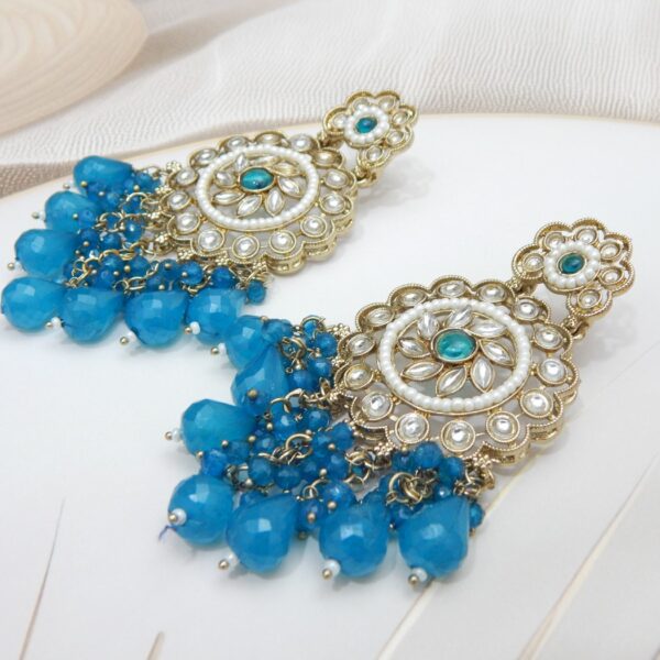 A pair of large, traditional dangle earrings with a central oval stone, intricate work, a halo of small pearls, and a fan of dangling blue colored beads.