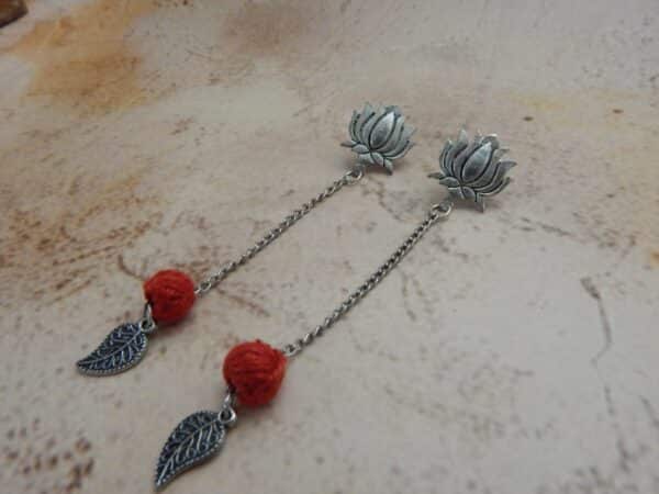 A pair of oxidized silver bohemian earrings featuring a lotus stud, a hanging red woven bead, and a leaf charm