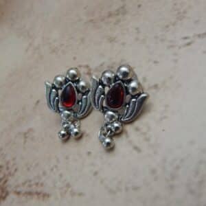 A pair of silver-toned oxidised stud earrings with a lotus flower design, a central red stone, and hanging beaded drops.