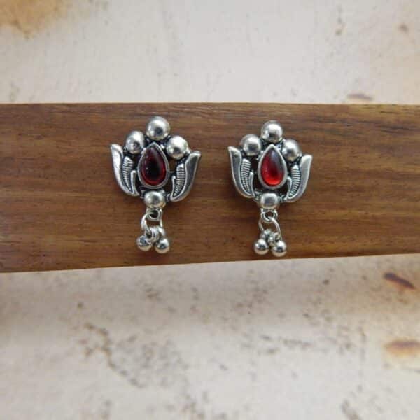 A pair of silver-toned oxidised stud earrings with a lotus flower design, a central red stone, and hanging beaded drops.
