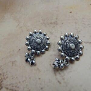 A pair of oxidized silver-toned earrings with a circular mandala-style stud and small jingle bells (ghungroos) dangling from the bottom