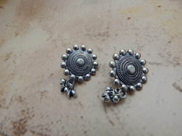 A pair of oxidized silver-toned earrings with a circular mandala-style stud and small jingle bells (ghungroos) dangling from the bottom