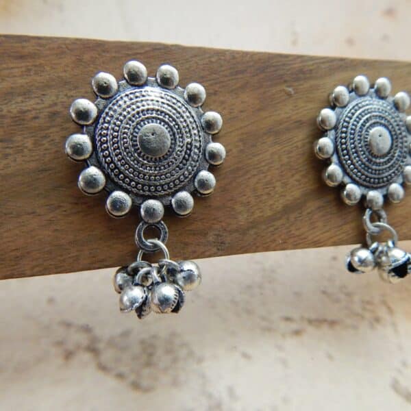 A pair of oxidized silver-toned earrings with a circular mandala-style stud and small jingle bells (ghungroos) dangling from the bottom