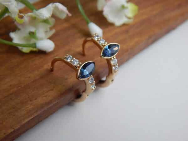 A pair of small, gold-toned huggie hoop earrings with a central dark blue, marquise-cut stone and side studs.