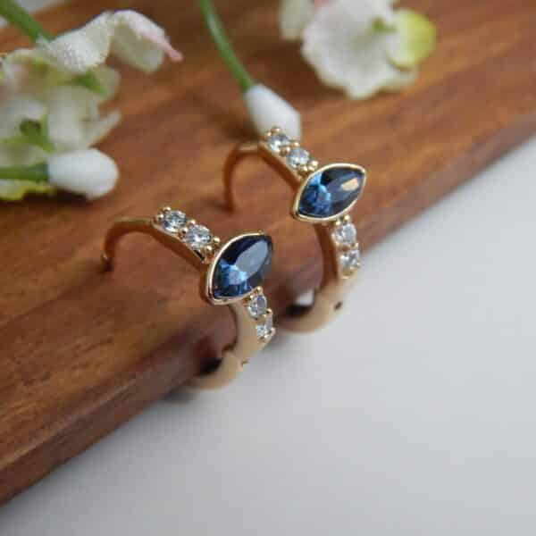 A pair of small, gold-toned huggie hoop earrings with a central dark blue, marquise-cut stone and side studs.