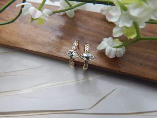 A pair of small, gold-toned huggie hoop earrings with a central clear, marquise-cut stone and side studs.