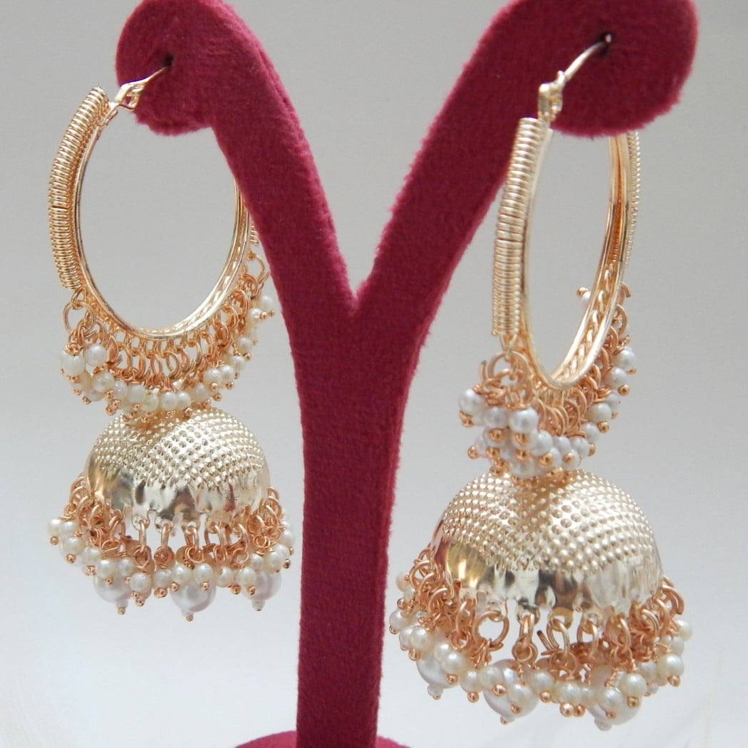 A pair of gold hoop jhumka earrings with white pearls, displayed on a red jewelry stand.