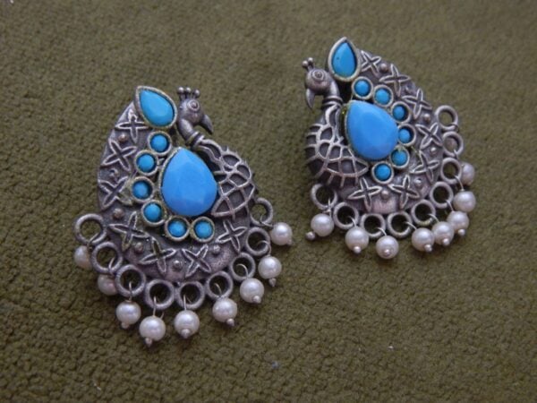 A pair of oxidized silver ethnic earrings with a peacock design, turquoise stones, and small pearl dangles.