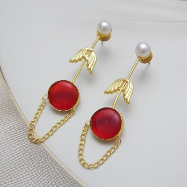 A pair of long gold-tone arrow-design earrings with a pearl stud, a round red colored stone, and a delicate chain loop.