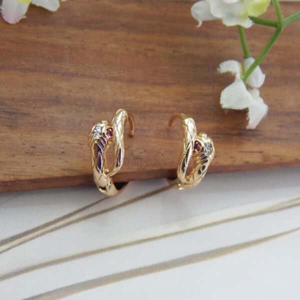 A pair of small, gold-toned huggie hoop earrings with a serpentine knot design and tiny red and white stones