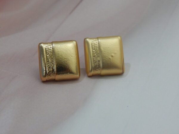 A pair of small, square, gold-toned stud earrings with a matte finish and a central ribbed texture.