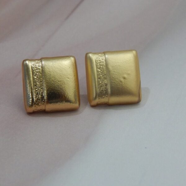 A pair of small, square, gold-toned stud earrings with a matte finish and a central ribbed texture.