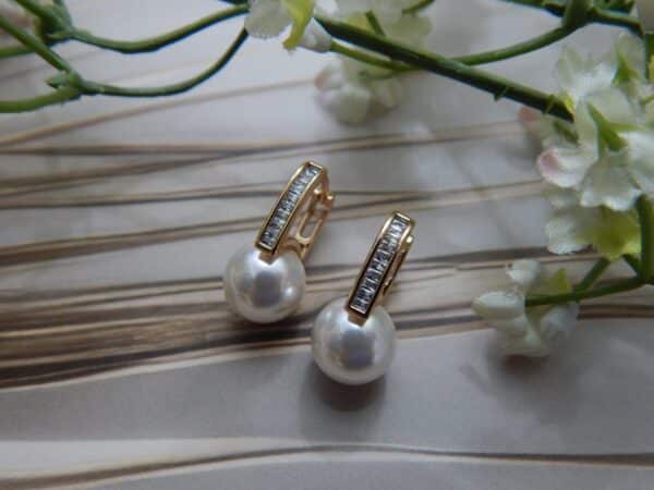 A pair of small, gold-toned huggie hoop earrings with a studded bar and a large white pearl drop