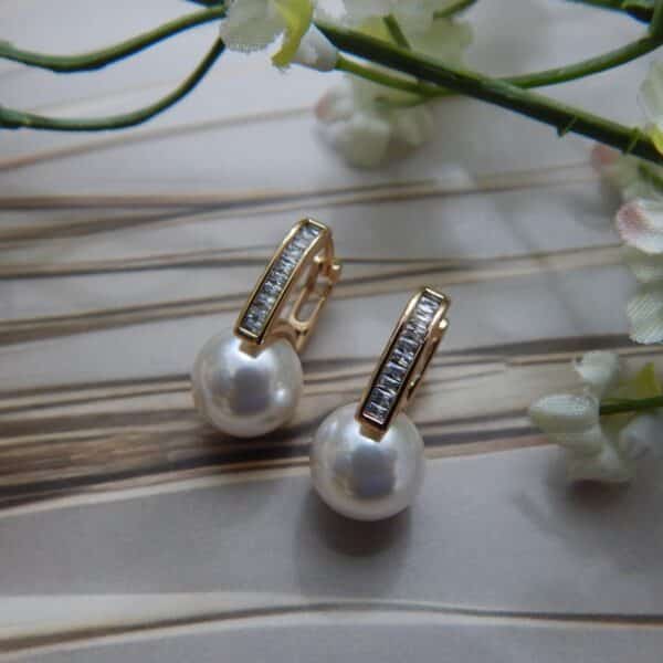 A pair of small, gold-toned huggie hoop earrings with a studded bar and a large white pearl drop