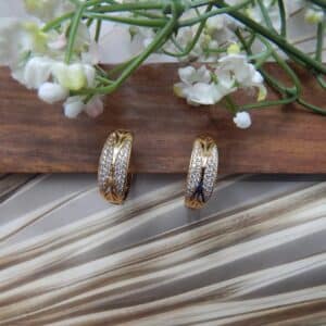 A pair of small, gold-toned huggie hoop earrings with a wide, wavy pattern of clear studs.