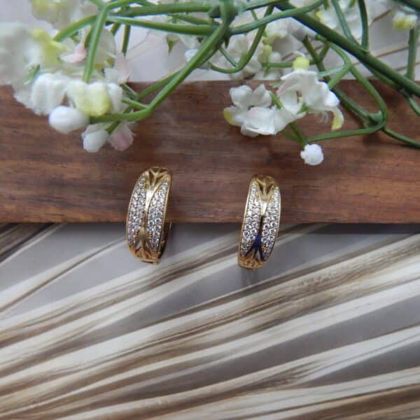 A pair of small, gold-toned huggie hoop earrings with a wide, wavy pattern of clear studs.