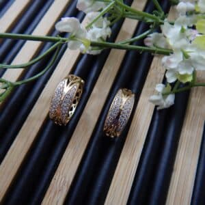 A pair of small, gold-toned huggie hoop earrings with a wide, wavy pattern of clear studs.
