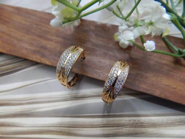 A pair of small, gold-toned huggie hoop earrings with a wide, wavy pattern of clear studs.