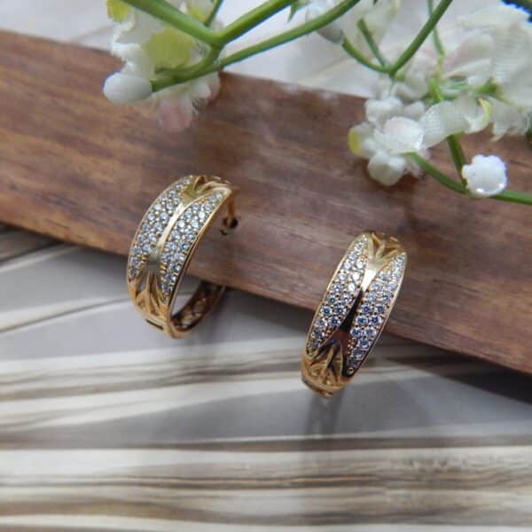 A pair of small, gold-toned huggie hoop earrings with a wide, wavy pattern of clear studs.