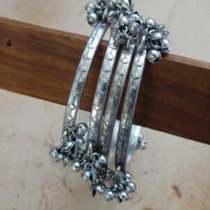 Close-up of four oxidized silver-toned bangles on stand with tribal etching and dangling ghungroo bells, ideal for traditional and college wear.