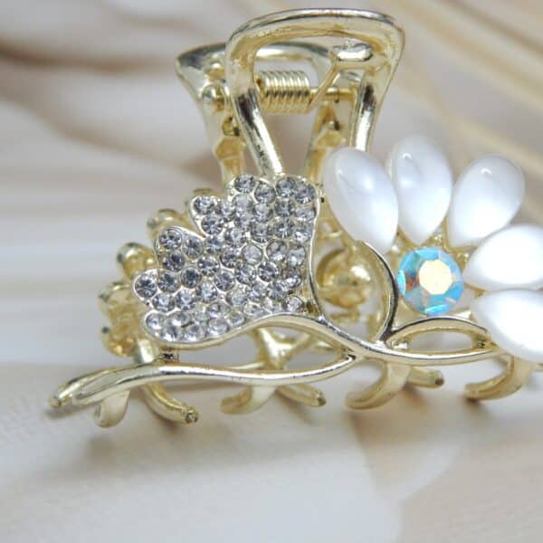 Small gold-toned hair claw clip with clear rhinestones and opalescent white/off-white cat-eye flower petals, elegant hair accessory for women.
