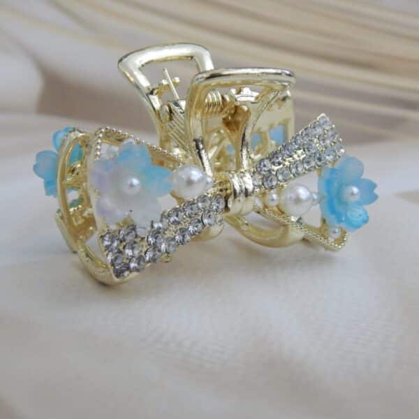 Small gold-toned hair claw clip with a crystal bow, pearl beads, and small blue resin flowers, elegant hair accessory for women.