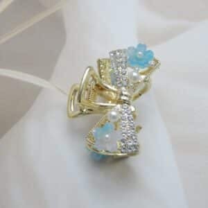 Small gold-toned hair claw clip with a crystal bow, pearl beads, and small blue resin flowers, elegant hair accessory for women.