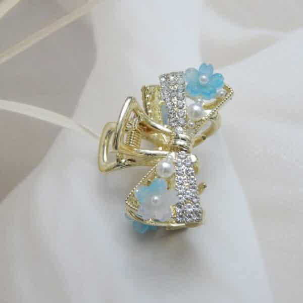 Small gold-toned hair claw clip with a crystal bow, pearl beads, and small blue resin flowers, elegant hair accessory for women.