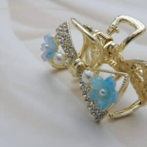 Small gold-toned hair claw clip with a crystal bow, pearl beads, and small blue resin flowers, elegant hair accessory for women.