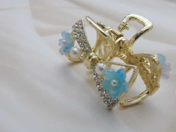 Small gold-toned hair claw clip with a crystal bow, pearl beads, and small blue resin flowers, elegant hair accessory for women.