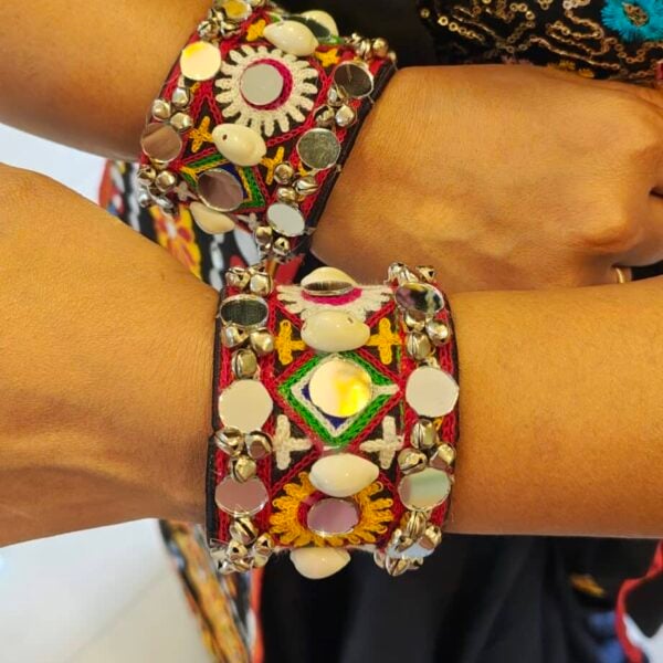 Red embroidered cuff bracelet with mirror discs, cowrie-style details and silver-finish bells worn on both wrists